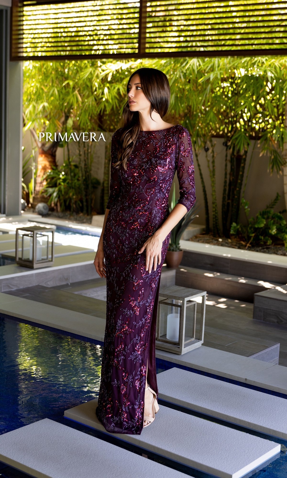 A woman in the Primavera 13116 Sleeved Long Beaded Formal Dress poses by an outdoor pool with modern decor and greenery in the background.